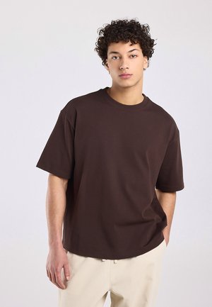 Jonge man met krullend haar die een loszittend donkerbruin kortemouwenhemd en lichtbeige broek draagt, staand tegen een effen achtergrond.