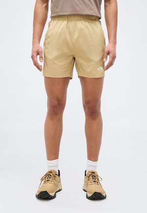 Muž oblečený v béžových šortkách, zodpovedajúce béžové turistické topánky s čiernymi podrážkami, biele členkové ponožky, stojí s uvoľnenými rukami po stranách.