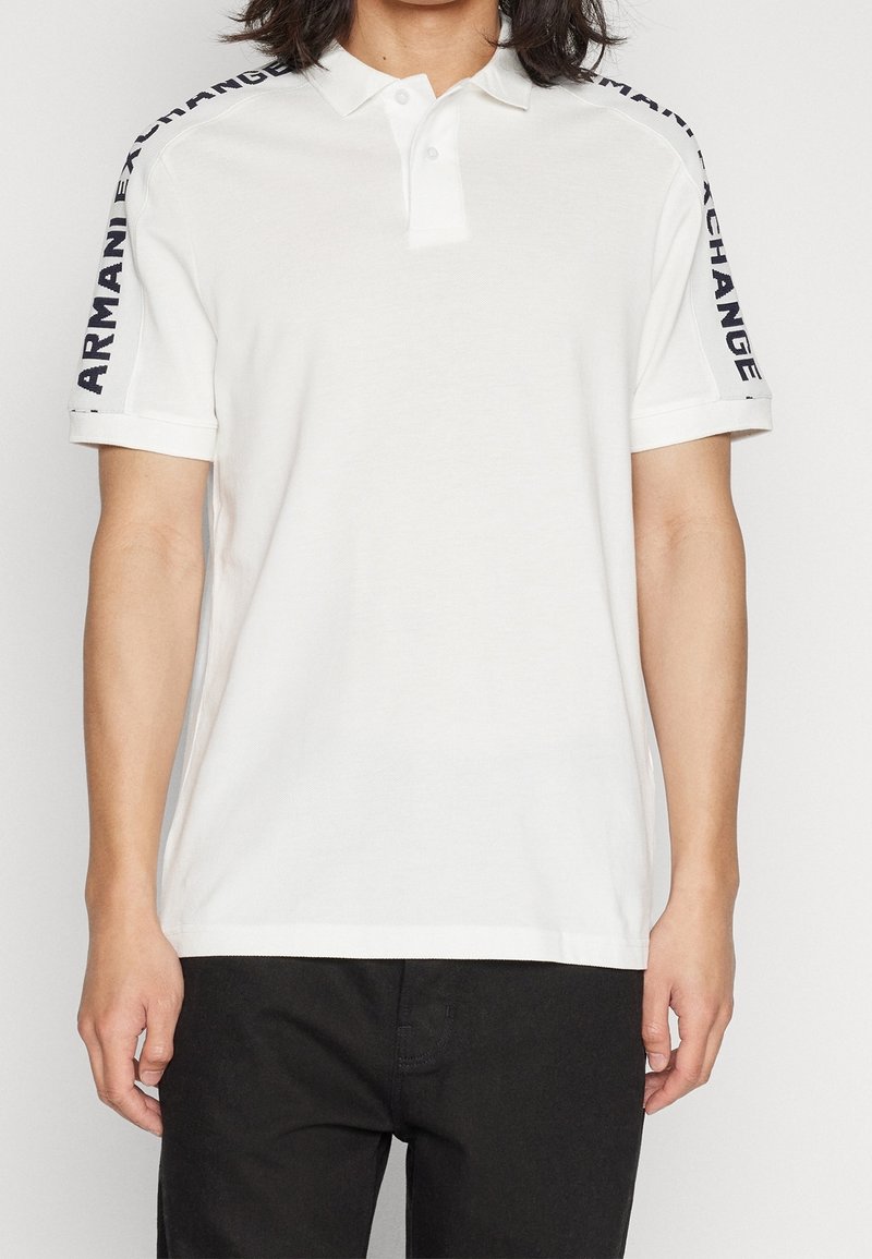 Polo blanc à manches courtes avec le texte noir "ARMANI EXCHANGE" sur les deux bandes d'épaule, porté avec un pantalon noir.