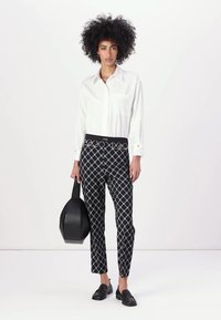 White long-sleeve shirt with chest pocket, paired with black patterned trousers featuring gold accents. Model holds a black bag.