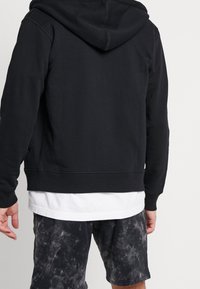 Felpa nera con cappuccio in tessuto morbido, dotata di cordino, polsini e vita a costine, abbinata a pantaloni corti scuri a fantasia.