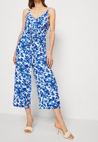 Donna che indossa un tuta senza maniche con stampa floreale blu e bianca, con pantaloni a gamba larga e vita legata, abbinata a sandali con tacco intrecciati color beige.