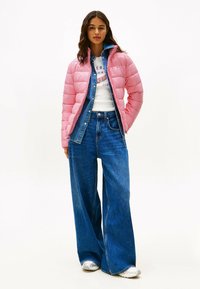 Lyserød puffjakke, denimskjorte, grafisk hvid t-shirt, brede blå jeans og sølvfarvede sneakers; stylet med langt, bølget hår.