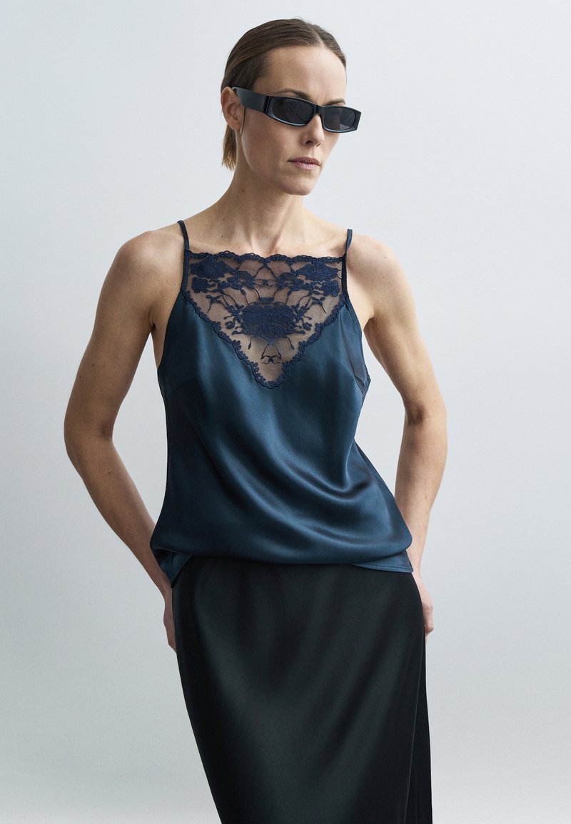 Tengerészkék szatén camisole csipkés nyakkivágással; vékony pántokkal és sima textúrával; fekete szoknyával párosítva, sötét napszemüveggel kiegészítve.