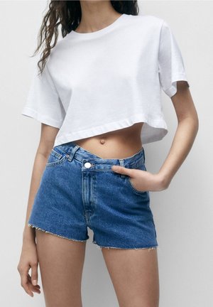 Vrouw draagt een wit cropped T-shirt en blauw hoogwater spijkershorts met rafelige zomen, staand met één hand in haar zak.