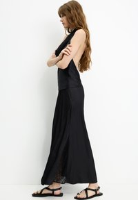 Schwarzes ärmelloses Kleid mit tiefem Rückenausschnitt, das eine fließende Silhouette und Spitzenakzente am Saum aufweist. Das Model trägt schwarze Sandalen.