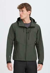 Homme portant une veste softshell à capuche vert fonc�é avec des poches zippées, des poignets ajustables et un pantalon noir, regardant vers sa gauche.