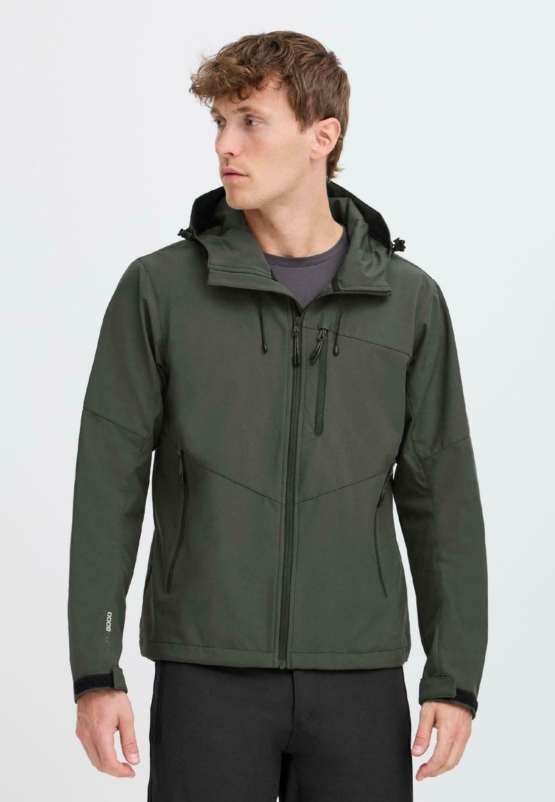 Homme portant une veste softshell à capuche vert foncé avec des poches zippées, des poignets ajustables et un pantalon noir, regardant vers sa gauche.