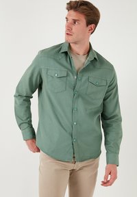 Cămașă verde cu nasturi, realizată dintr-un material texturat, având două buzunare la piept și nasturi tip clapetă, combinată cu pantaloni bej deschis.