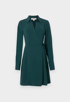 Rochie verde cu mâneci lungi, guler și un nod pe lateral. Dispune de un buzunar pe piept și o textură netedă cu o fustă vaporoasă.