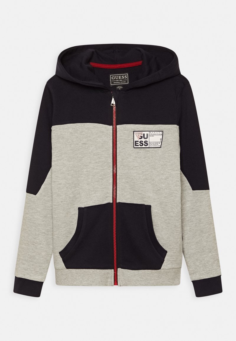 Sweatshirt à capuche zippé en gris et bleu marine, avec une fermeture éclair rouge, des poches avant et un patch logo sur la poitrine. Matière en mélange de coton.