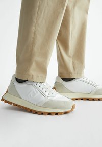 Sneakers atletici bianchi con inserti in suede grigio, suole di gomma testurizzate e un design affusolato. Indossati con pantaloni beige a gamba affusolata.