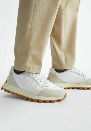 Sneakers atletici bianchi con inserti in suede grigio, suole di gomma testurizzate e un design affusolato. Indossati con pantaloni beige a gamba affusolata.
