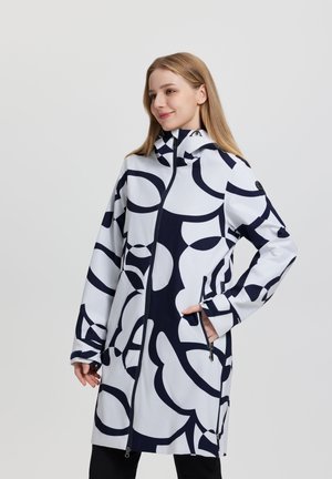 Frau trägt eine weiße lange Jacke mit großen schwarzen abstrakten Mustern, steht mit einer Hand in der Tasche vor einem schlichten hellen Hintergrund.