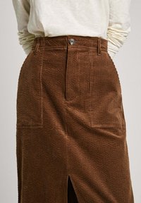 Falda de pana marrón con una cintura ajustada, detalles de bolsillos delanteros y una abertura frontal. La tela tiene una textura acanalada y un cierre de botón.