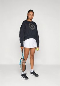 Granatowa bluza z kapturem ze złotym logo, biała tenisowa spódniczka, czarne trampki, trzyma rakietę tenisową i piłkę. Wnętrze, neutralne tło.