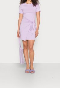 Lavendelfärgad t-shirtklänning med knutdetalj vid midjan, korta ärmar, matchad med en lila meshväska och flätade sandaletter. Slät tygtextur.
