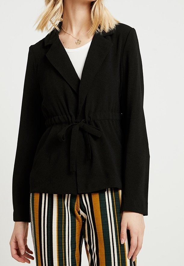 Femme portant un blazer noir avec une taille nouée sur un haut blanc et un pantalon rayé de lignes verticales vertes, jaunes, noires et blanches.