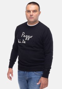 Maglione nero lavorato a maglia con la scritta "Pazzo di Tequila" ricamata in bianco. Maniche a costine e orlo, abbinato a jeans in denim blu.