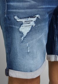 Shorts de denim azul con dobladillo vuelto y un desgarro deshilachado prominente en el muslo que muestra hilos blancos debajo.