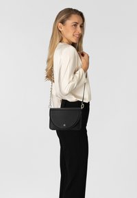 Borsa a tracolla in pelle nera con finitura morbida e strutturata, patta curva, tracolla a catena argentata e dettaglio logo discreto.