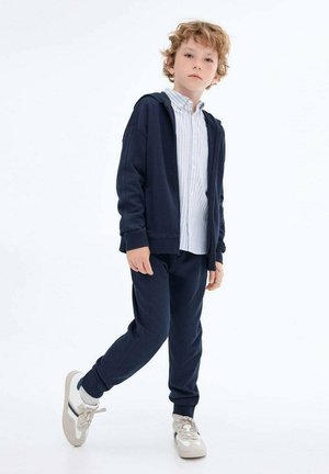 Felpa blu navy con cappuccio, abbinata a pantaloni joggers coordinati. Sotto, una camicia a righe blu chiaro con bottoni. Sneakers bianche con dettagli grigi.