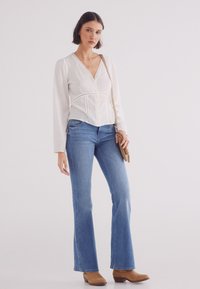 Weiße Langarmbluse mit strukturiertem Detail, kombiniert mit hellblauen Schlagjeans und braunen Ankle Boots. Das Model hält eine kleine braune Umhängetasche.