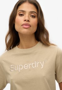 Beige katoenen t-shirt met een zacht, geborduurd "Superdry" logo in een iets donkerdere beige tint. Ronde hals en korte mouwen.