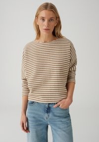 Maglione a righe in beige e bianco; ampia scollatura a barca, spalle scese e vestibilità rilassata. Abbinato a jeans in denim azzurro chiaro.