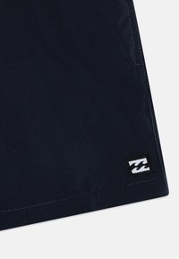 Jūras spēku audums ar gludu tekstūru, kuram ir melnbalts viļņu logotipa plāksteris un kontrastējoša šuvuma apdare pa malām.