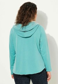 Turkoosi huppari, jossa on pehmeä rakenne, rento istuvuus ja viimeistelemätön helma. Yhdistetty tummiin, kuvioituihin housuihin.