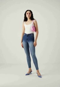 Modelo vistiendo una camiseta blanca sin mangas, jeans ajustados azules y zapatos de tacón ligeros azules, sosteniendo un pequeño bolso rosa. Fondo simple.