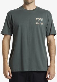 Camiseta de algodón de color verde oscuro con mangas cortas, que presenta un pequeño diseño gráfico multicolor en la parte izquierda del pecho. Corte estándar.