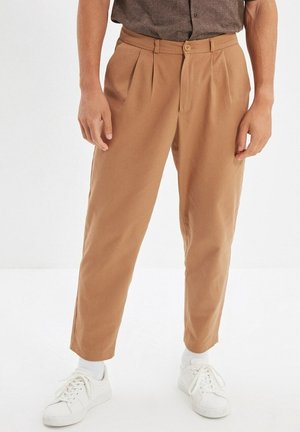 Homme portant un pantalon court marron clair associé à des baskets blanches et des chaussettes blanches, debout devant un fond blanc uni.