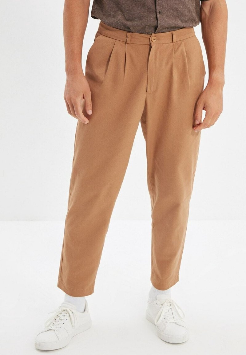 Homme portant un pantalon court marron clair associé à des baskets blanches et des chaussettes blanches, debout devant un fond blanc uni.
