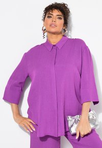 Camicia a maniche corte di colore viola, realizzata in un tessuto strutturato. Il modello tiene una pochette argentata, esposta su uno sfondo bianco.