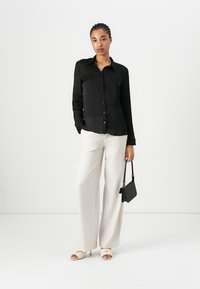 Svart skjorte med knapper, lange ermer, linstoff. Hvite vide bukser, glatt tekstur. Lyse sandaler med minimalistisk design. Svart håndveske.
