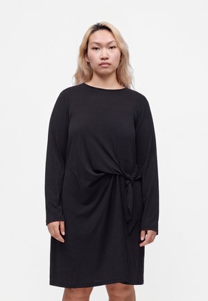Kvinde med lyst blond hår iført en langærmet sort kjole med bindebånd foran mod en ensfarvet hvid baggrund.