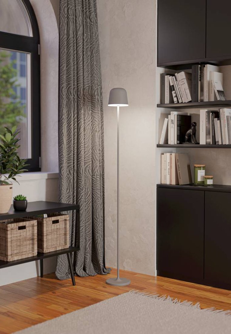 Hohe, schlanke Stehlampe mit einem grauen Schirm und einem Metallfuß. Umgebende Elemente umfassen einen Tisch aus dunklem Holz, geflochtene Körbe und gemusterte Vorhänge.