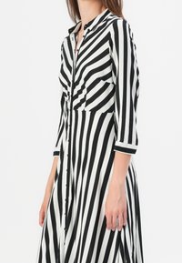 Robe chemise noire et blanche à rayures avec un devant boutonné, un col et des manches trois-quarts. Elle présente deux poches poitrine et une coupe fluide.