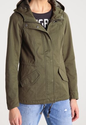 Vrouw draagt een olijfgroene gewatteerde jas met capuchon, klepzakken en een ritssluiting aan de voorkant, over een zwart shirt en gescheurde blauwe spijkerbroek.