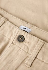 Beige bomullsbukser med knappestenging, merke med teksten "TWO SOON" og synlig sømm detalj langs midjen.