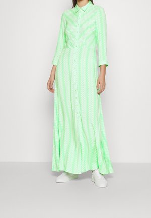 Femme portant une robe maxi à manches longues, boutonnée sur le devant, de couleur vert néon avec un motif chevron blanc, et des baskets blanches, debout devant un fond blanc.