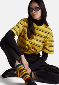 Žluté tričko s černými proužky a logem "Stranger Things". Nosit přes černé tričko s dlouhým rukávem, doplněno barevnými pruhovanými ponožkami.