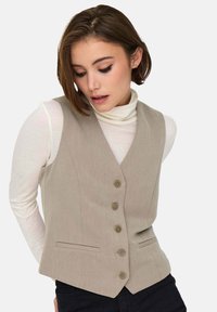 Gilet beige su misura con cinque bottoni, scollatura a V pronunciata e due tasche frontali; indossato sopra un dolcevita bianco. Texture del tessuto liscia.