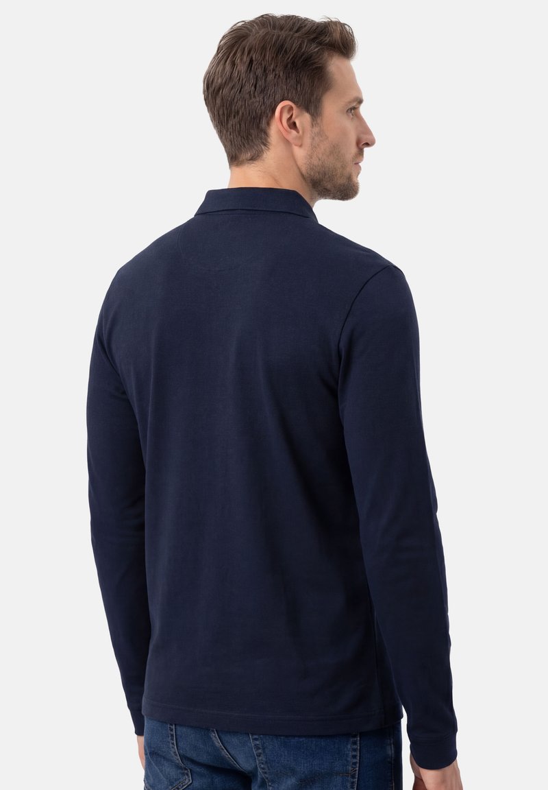Homme vu de dos portant un polo à manches longues bleu marine et un jean bleu, sur un fond clair uni.