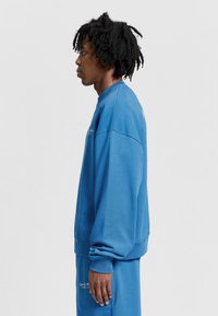 Niebieski sweter z okrągłym dekoltem, wykonany z miękkiego materiału, o luźnym kroju, z żebrowanymi mankietami i haftowanym logo na plecach. Gładka faktura.