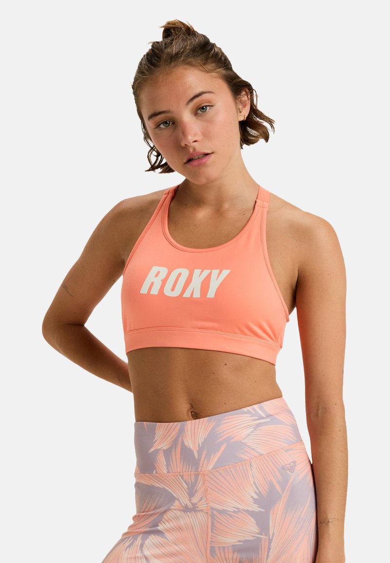Roxy sportbeha in koraal met een witte logo, voorzien van een racerback ontwerp. Gelaagd stof en aansluitende pasvorm. Gecombineerd met een gepatternde legging.