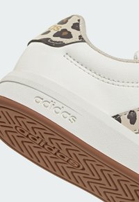 White sneaker with a textured upper, featuring a leopard print heel tab and embossed "adidas" logo. Solid rubber sole with a zigzag pattern.