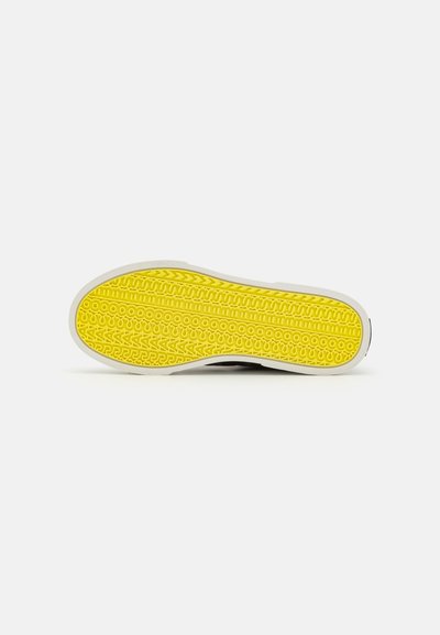 Shoe sole with a textured yellow rubber surface featuring circular and zigzag patterns, bordered by white material. Black upper edge visible.
